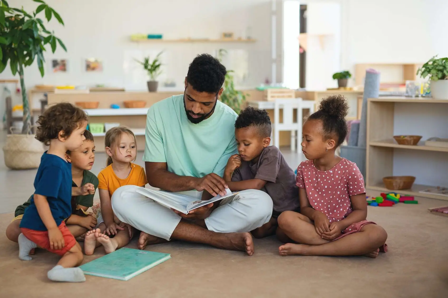 Nursery school children with man teacher sitting on floor in classroom, reading fairy tale book.